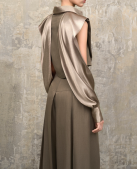 A model wearing a sleeveless, draped satin top and matching wide-leg trousers in a taupe-brown color, standing against a textured gray concrete wall.