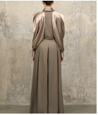 A model shown from the back wearing a floor-length taupe dress with draped sleeves, standing against a textured concrete wall.