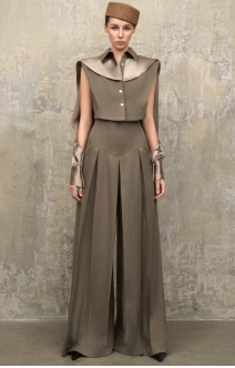 A model wearing a structured, sleeveless taupe-brown dress with a high collar, button front, and wide legs, paired with a matching brown hat, standing against a textured gray wall.
